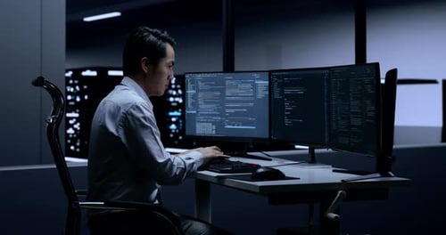 Vista lateral de um programador asiático do sexo masculino escrevendo código por desktops na sala de servidores