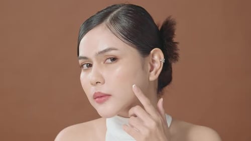 Young beautiful woman is checking her skin and popping pimple over brown background