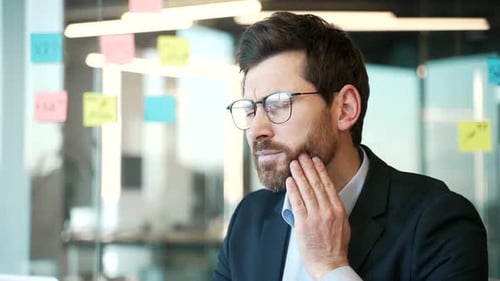 Upset businessman in a formal suit suffering from toothache while sitting at workplace in business