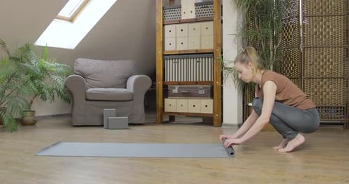 Blonde woman rolls up her yoga mat in her home studio.