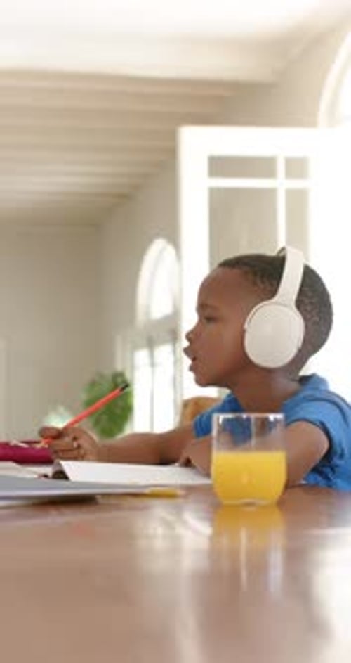Vertical video: Headphones playing audio, causing kid to write with red pencil and gesture at table