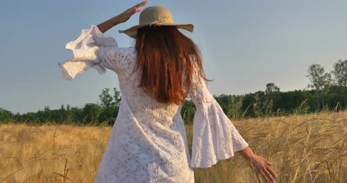 On a sunny day, a woman is walking along a wheat field with a white dress, in a straw hat on a nat