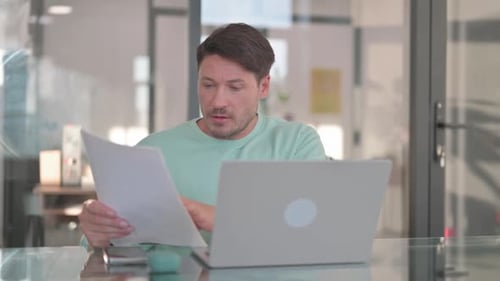 Man Reads Papers While Working on Laptop in Office