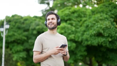 Smiling sporty man walking with wireless headphones using browsing mobile phone on urban city park.
