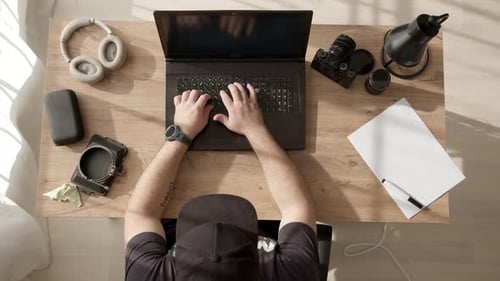 Freelancer masculino irreconhecível com boné preto está sentado à mesa e digitando no laptop em casa