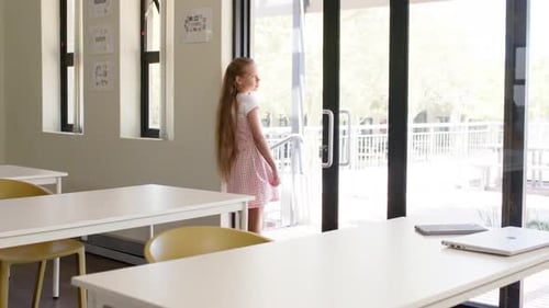Preteen girl in gingham dress walking from glass doors to table with laptop, notebook, copy space
