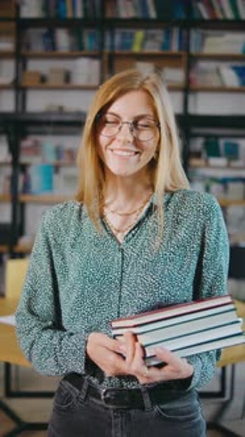 A Young Woman with Glasses in a Library Smiling Holding Books and Browsing Shelves Woman Holding a