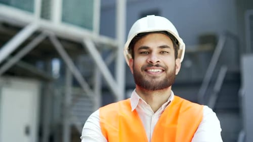 Portrait of smiling professional engineer wearing safety helmet and vest standing at factory.