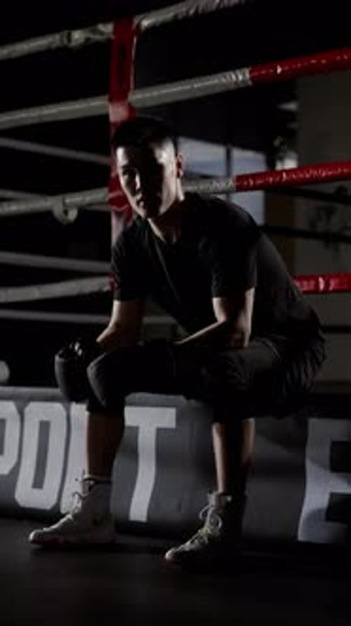 Focused Young Adult Boxer Sits in Boxing Ring