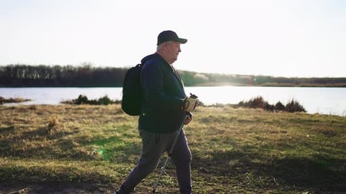 Active Handsome Elderly Man with Nordic Walking Sticks Walks Forward Through Along the Lake at