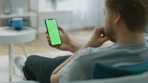 Young Man at Home Uses Green Mock-up Screen Smartphone. She's Sitting On a Couch in His Cozy Living