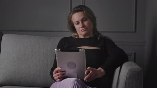 A Woman is Enjoying a Leisurely Moment on Her Cozy Home Sofa While Using a Tablet Device to Unwind