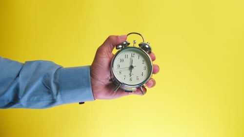 Hand Holding Silver Alarm Clock Against Yellow Background