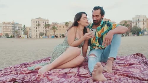 Young couple uses mobile phone while sitting on the beach on buildings background