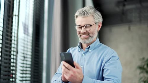 Mature gray haired bearded businessman browsing smartphone at workplace in business office. Senior