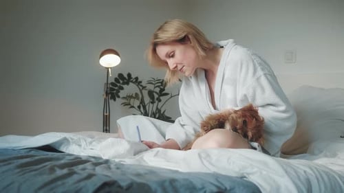 Woman Writes on Bed with Small, Brown Dog