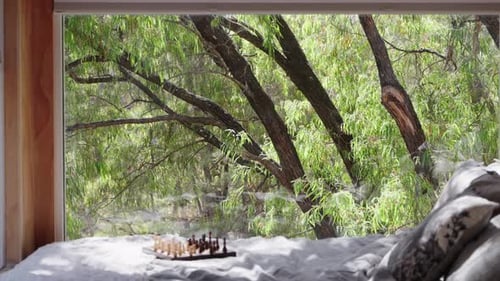 Chess board sitting a top of tiny cabin bed with large window out to the forest