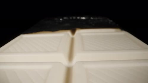 A white chocolate bar, partially unwrapped from the foil, on a black background. Dolly macro
