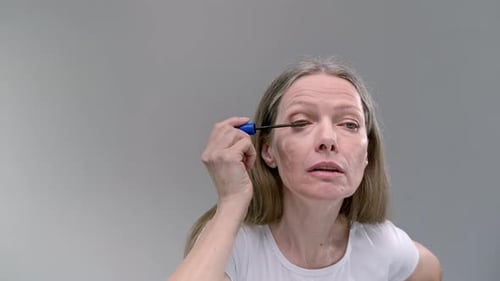 Senior Lady Applies Mascara Onto Eyelashes with Brush Looking in Mirror on Light Grey Background