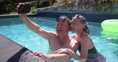 Casal sênior na piscina, homem estendendo o telefone tirando selfie, mulher em traje de banho inclinado verde para trás
