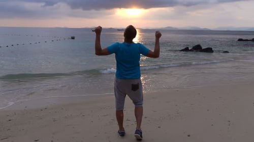 Fröhlicher junger Mann hebt am exotischen Strand bei Sonnenuntergang in Superzeitlupe die Arme zum Himmel