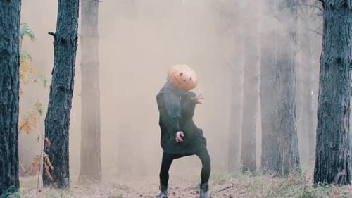 A Funny Girl in Black Clothes with a Large Pumpkin Head Dances in the Orange Smoke in the Open Air