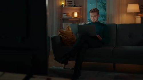 In the Evening Handsome Young Man at Home Sitting on a Couch in the Living Room Works on a Laptop C