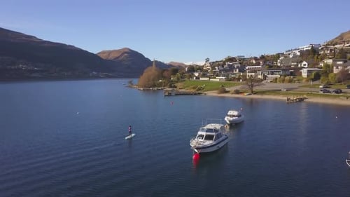 Paddleboarder in paddling in a blue bay next to moored boats. Drone panning shot