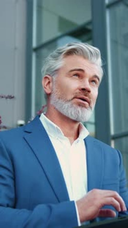 Mature Businessman Working Thoughtfully on a Laptop Outdoors with a Modern Glass Building in the