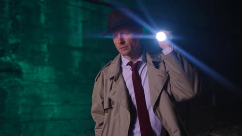 Stylish Portrait of a Searching Detective in a Raincoat and Hat with a Flashlight Looking for Clues