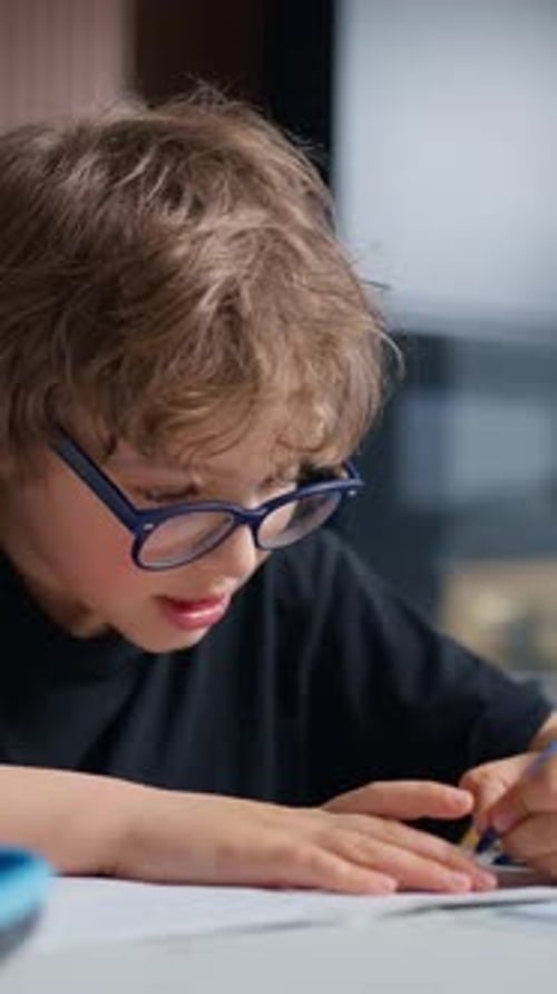Young Boy with Curly Hair and Glasses Focused Intently on Writing or Drawing on Paper with a Pencil
