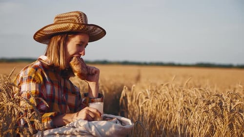 Young Girl Farmer in Sun Hat Eats Loaf of Bread Biting It Drinking Milk Sits on Wheat Field with