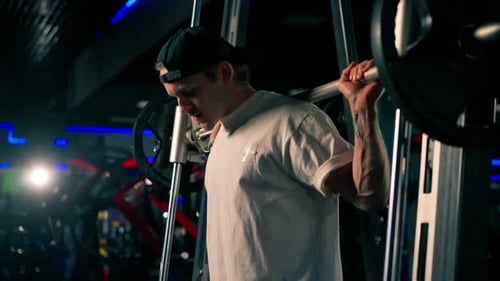 Close Up in the Gym Young Handsome Guy in a Tshirt and a Cap Shakes His Back with a Barbell with
