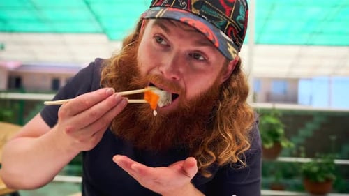 Close up bearded man eats Japanese sushi rolls with chopsticks in a street cafe.