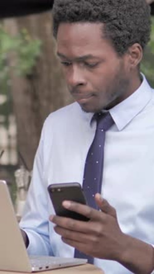 African Businessman Using Smartphone and Laptop in Outdoor Cafe, Vertical Video