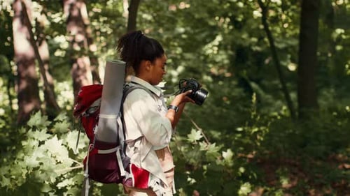 Une femme à la peau foncée se promène dans la forêt tout en tenant un appareil photo et un sac à dos pendant la journée
