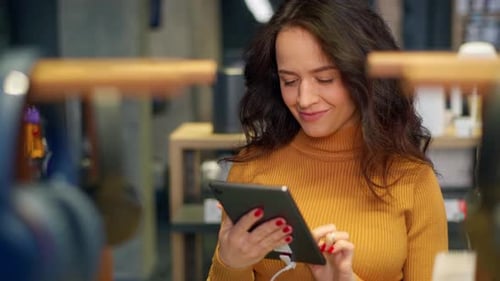 Attractive young girl with curly hair in an orange sweater chooses and tests a tablet in a store.