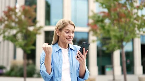 Excited woman is happy about great news she read on phone while standing on street near a modern