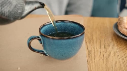Teekanne zum Aufbrühen und Servieren von Tee, Eingießen von grünem Tee in eine blaue Keramiktasse auf einem hölzernen Cafétisch Zwei