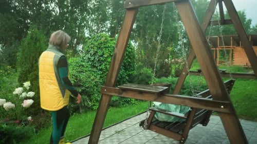 Woman in Yellow and Green Jacket Walks Toward Swing Bench with Mint Bag in Garden
