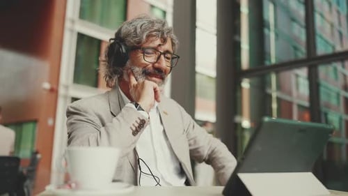 Mature businessman and headphones, sits in an outdoor cafe and works using tablet