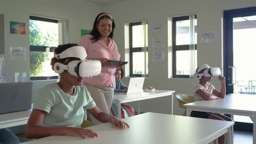 African teacher walking in class urging teenage female donning VR headset reaching exploring lesson
