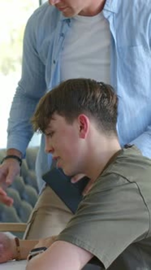 Vertical video: Leaning adult showing dark device guiding teen writing notebook at home