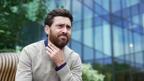 Sick businessman suffering from a sore throat while sitting on a bench on the street near business