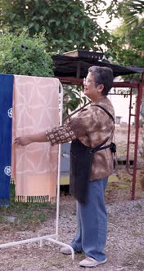 Senior Asian woman artisan hangs fresh indigo dyed textile on outdoor rack, smooths edges, checks