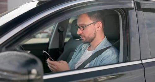 Young Caucasian Man Sits in a Car on a Parking Lot and Typing Message on a Smart Phone