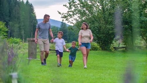 Young Family Walking Along Grass In Park Beside River Holding Hands And Waving To Someone. Slow Mot