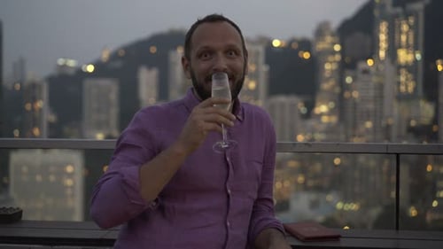 Handsome man smiling and raising a toast with cocktail in luxurious city skybar