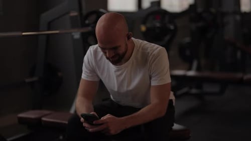 Man Using Phone and Earbuds Resting After Fitness Workout in Gym