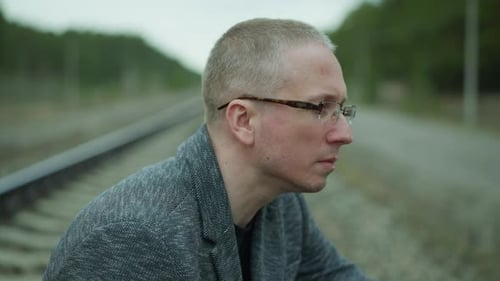 Contemplative Man in Glasses Sitting By Railway Track in Serene Forest